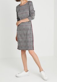 Robe à carreaux à manches longues, avec un motif noir, blanc et rouge. Rayures sur les côtés en rouge, associées à des baskets blanches.