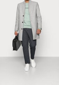 Cappotto grigio chiaro con chiusura a bottoni, felpa verde menta, pantaloni grigio scuro e sneakers bianche. Porta una valigetta in pelle nera.