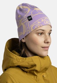 Purple knitted beanie with abstract beige patterns, featuring a black logo tag. Worn with a mustard yellow jacket, showing smooth texture and fit.