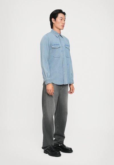Lys blå denimskjorte med lange ermer, to brystlommer, knappestolpe, brukt med mørkegrå slengjeans og svarte sko.