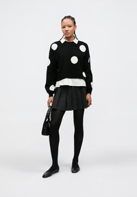 Μαύρο πουλόβερ με άσπρες πουά, φορεμένο πάνω από λευκό πουκάμισο με γιακά· πλισέ μαύρη φούστα, μαύρο καλσόν και φλατ παπούτσια. Κρατάει μαύρη τσάντα.