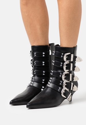Black leather ankle boots with pointed toes, featuring decorative studs and four silver buckles along the shaft. Textured and sleek design.