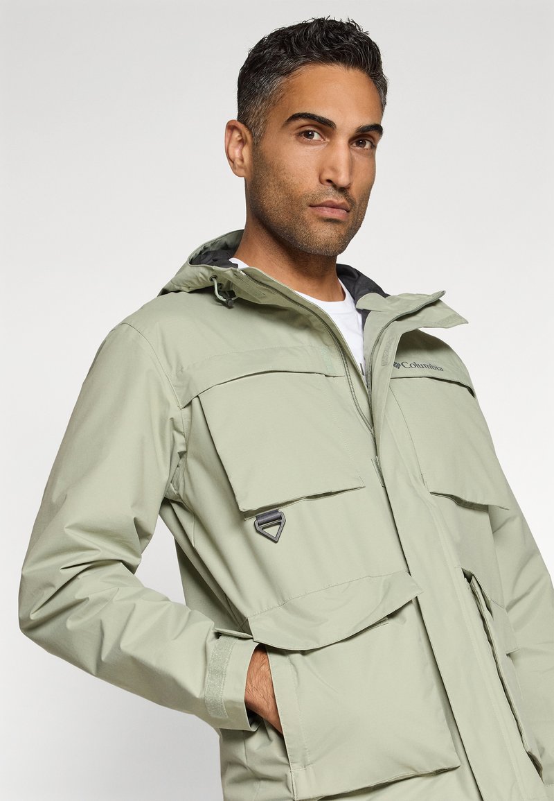 Jasnozielona wodoodporna kurtka z zamkiem z przodu, dużymi kieszeniami z klapami i kapturem. Posiada kontrastową czarną podszewkę oraz sprzęt zamka.