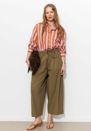 Femme debout contre un mur blanc, portant une chemise rayée orange, un pantalon large olive, des sandales marron clair, tenant une pochette marron à franges.