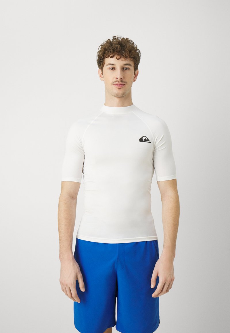 Hvit kortermet rashguard med rund hals, med en liten svart logo på brystet, kombinert med blå svømshorts.