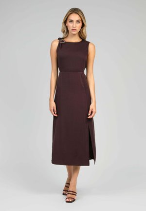 Femme blonde debout portant une robe midi marron foncé sans manches avec une boucle à l'épaule et une fente latérale, associée à des sandales marron à fines lanières.