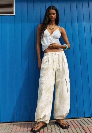 Fiatal nő hosszú sötét hajjal, fehér ujjatlan felsőt, krémszínű cargó nadrágot, szandált és türkiz karkötőt viselve, áll egy kék fal előtt.