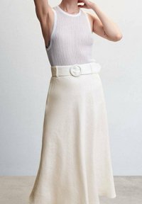 Nainen, jolla on ylleen puettu ribbattu valkoinen hihaton toppi, joka on työnnetty korkeavyötäröisen kermaisen hameen sisään. Hameessa on leveä vyö ja pyöreä solki.