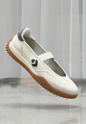 Hvid slip-on sko med brun gummisål, sort stjerne og chevron-logo på siden samt sort hælpude, placeret foran gråt gulv og baggrund med gardin.