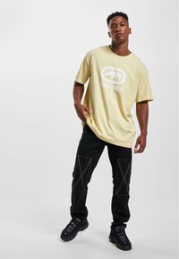 Lys gul oversized t-shirt med en hvid logo, parret med sorte bukser med kontrasterende hvid syning og en afslappet pasform. Sorte sneakers.
