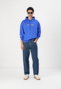Sudadera con capucha azul y bolsillo frontal, con el logotipo amarillo "RED BULL RACING", combinada con unos jeans de corte relajado y zapatillas blancas.