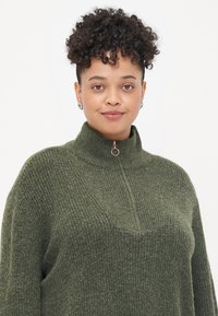 Suéter de punto texturizado verde con un alto cuello y detalle de media cremallera. Presenta un corte relajado y un patrón acanalado en toda su superficie.
