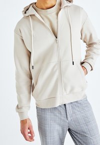 Felpa beige con zip e cordini, tessuto testurizzato, due tasche frontali. Abbinata a pantaloni a quadri grigi chiari con accenti blu.