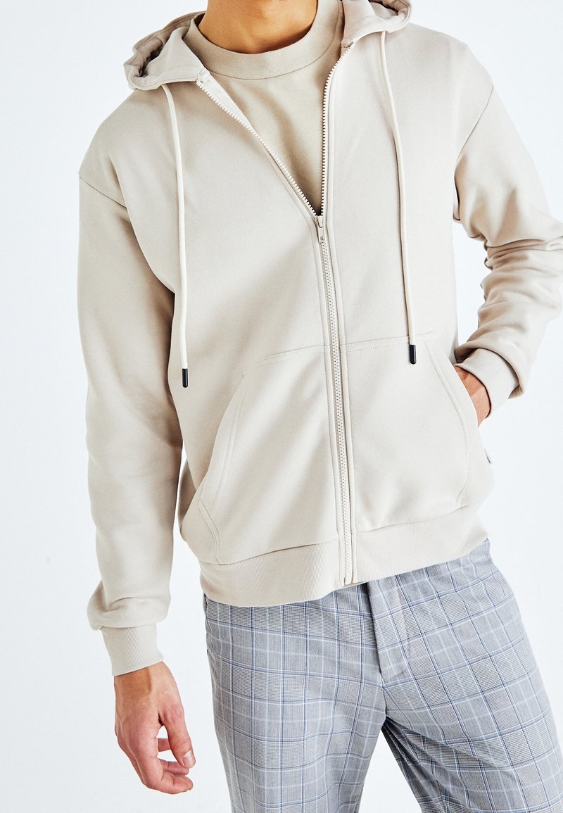 Felpa beige con zip e cordini, tessuto testurizzato, due tasche frontali. Abbinata a pantaloni a quadri grigi chiari con accenti blu.