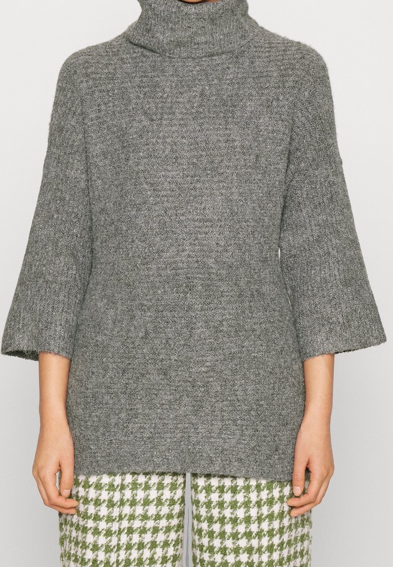 Pull à col roulé en tricot gris texturé avec des manches larges trois-quarts, associé à un pantalon à motif pied-de-poule vert et blanc.