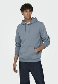 Lichtgrijze hoodie van zacht materiaal, met een voorzak, trekkoord capuchon en geribbelde manchetten. Gecombineerd met donkere spijkerbroek.