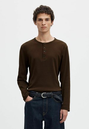 Jeune homme aux cheveux bouclés portant une chemise henley marron à manches longues, un jean foncé et une ceinture noire tressée, debout avec une main dans la poche.