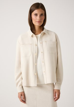 Vrouw in een off-white button-up jack met twee borstzakken over een fluffy witte trui en bijpassende rok tegen een effen achtergrond.