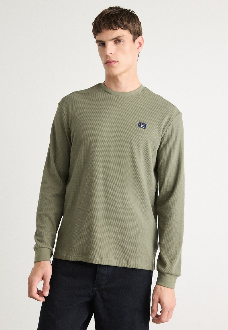 Olijfgroene langarmsg T-shirt van een gestructureerde ribstof, met een klein marineblauw logopatch op de borst. Gecombineerd met donkere jeans.