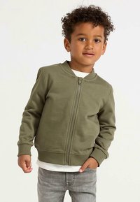 Olive green bomber jacket with a zip front, ribbed cuffs, and collar, paired with a white shirt and gray jeans.