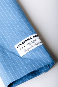 Ύφασμα με ανοιχτό γαλάζιο ρίγες και λείο υφή, με μια λευκή ετικέτα που αναγράφει "Colourful Rebel, Ready to Wear, Designed in Amsterdam."