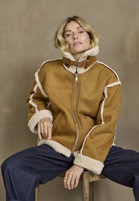 Bruine nubuck leren jas met schapenvacht voering, ritssluiting en een hoge kraag. Heeft contrasterende stofranden en een losse pasvorm.