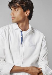 Witte overhemd met een klassieke kraag, met een geborduurd logo op de borst en een contrasterend blauw label aan de hals.