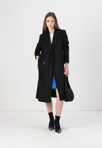 Cappotto nero doppiopetto con spalle rotonde, spacchi laterali e vestibilità strutturata. Indossato sopra un top blu, abbinato a una borsa nera texturizzata.