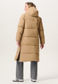 Vrouw in een tan lange gewatteerde jas met capuchon, zijsplitsingen, grijze wijde jeans en witte sneakers met zwarte strepen, staand met de rug naar de camera.