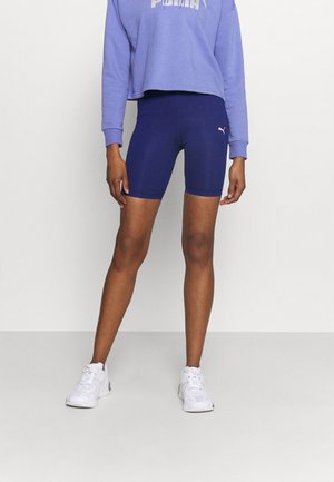 Sweat-shirt violet avec un design court, associé à un short de cyclisme bleu marine avec un petit logo. Des baskets blanches complètent la tenue.