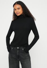 Czarna, gładka, długorękawowa bluzka z golfem o dopasowanym kroju, zestawiona z ciemnoszarymi, wysokim stanem jeansami, ręce w kieszeniach.