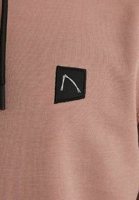 Felpa rosa chiaro con tessuto texturizzato, presenta una toppa quadrata nera con un design astratto bianco e una zip nera sul colletto.