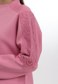 Rosa plagg med glatt stoff og intrikate blonde detaljer på ermene. Blonden har et blomstermønster, som gir tekstur og kontrast.