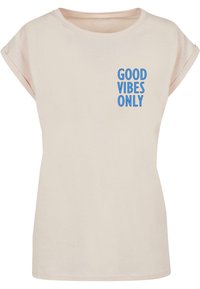 Helles beiges T-Shirt mit aufgerollten Ärmeln, rundem Halsausschnitt und blauer Schrift auf der linken Seite, die "GOOD VIBES ONLY" liest. Weiches Baumwollmaterial.