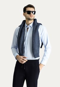 Camicia azzurro chiaro con un maglione grigio a coste drappeggiato sulle spalle. Pantaloni neri e occhiali da sole completano l'outfit.