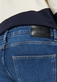 Tamne traperice tamnoplave srednje pranje, s kožnom etiketom "Marc O'Polo DENIM" na stražnjem pojasu.