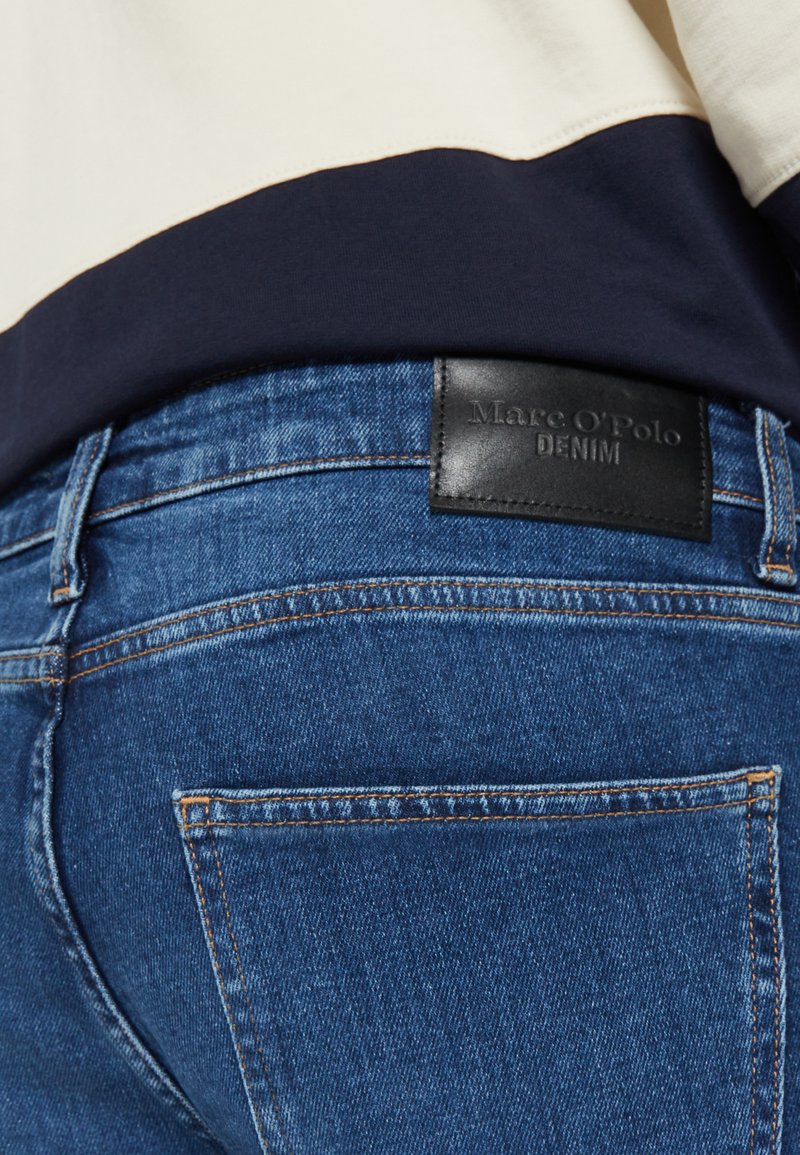 Jean en denim bleu foncé avec un lavage moyen, comportant une étiquette en cuir portant la mention "Marc O'Polo DENIM" à l'arrière de la taille.