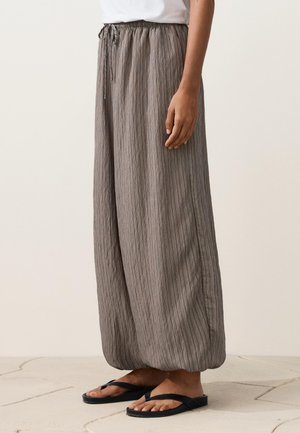 Persona che indossa pantaloni ampi color taupe, stropicciati, con vita elastica e infradito nere, in piedi su un pavimento chiaro in piastrelle.