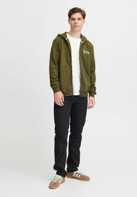 Olivengrønn zip-up hettegenser med frontlommer, kombinert med en hvit skjorte og sorte jeans. Brukt med grå joggesko med hvite snøringer.