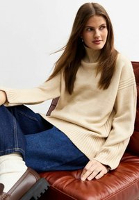 Suéter de cuello alto beige con dobladillo y mangas acanaladas, combinado con jeans de mezclilla oscura y zapatos marrones, sentado en una silla de cuero marrón.