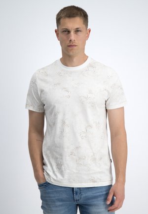 Ung mand med kort hår iført en hvid t-shirt med blomsterprint og blå jeans, stående med den ene hånd i lommen.