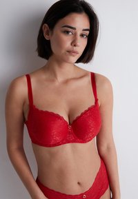 Soutien-gorge en dentelle rouge avec armatures, bords festonnés et un petit accent doré au centre. Bretelles réglables et texture lisse.