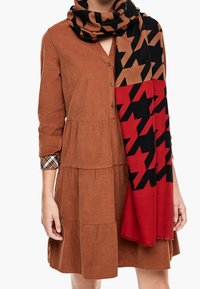 Robe boutonnée brune avec un design à étages, accompagnée d'un foulard polyvalent à motif pied-de-poule en noir, rouge et beige.