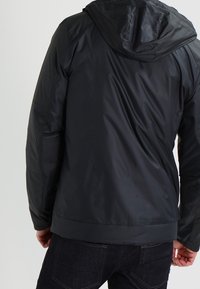 Mörk, vattentät jacka med huva och slät textur. Har elastiska muddar och en figursydd nederkant, vilket visar en elegant och modern design.