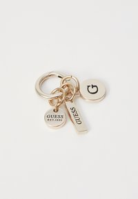 Zlatý klíčenka se třemi přívěsky: jedním kruhovým s nápisem "GUESS EST.1981", jedním obdélníkovým s nápisem "GUESS" a jedním kruhovým s logem "G".