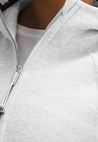 Sweat à capuche gris clair avec fermeture éclair, doté d'une texture douce, comprenant une capuche et une fermeture éclair argentée. Le tissu semble lisse sans motifs visibles.