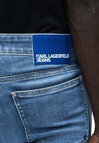 Κοντινή λήψη μπλε τζιν με μπλε ετικέτα που γράφει "KARL LAGERFELD JEANS" στη μέση, φορεμένα με μαύρο τοπ.