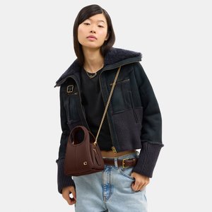 Fiatal nő sötét műszőrméből készült dzsekit és világoskék farmert visel, vállán kis barna kézitáskát arany lánc pánttal.