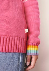 Roze gebreide trui met geribde manchetten en regenboogstrepen. Etiket aan de zijkant met de tekst "Little Bird." Denim jeans die gedeeltelijk zichtbaar zijn.