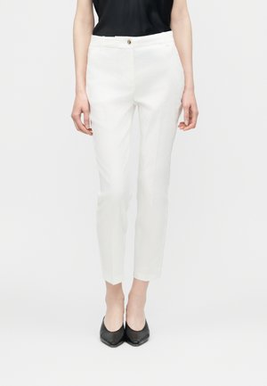 Naine, kes kannab valgeid sirgelõikelisi pükse esitaskute ja nööpidega, mida kannab koos mustade terava ninaga kingade ja musta topiga.